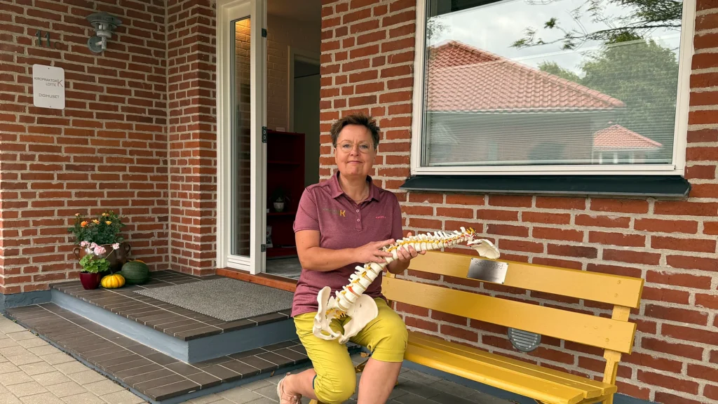 Kiropraktor Lotte Krogh ved den åbne indgangsdør til klinikken med en model af rygsøjlen, siddende ved den gule bænk på trappestenen