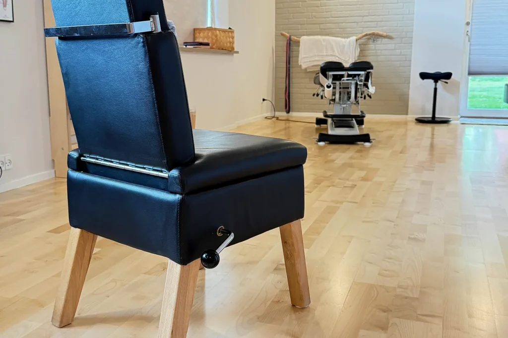 Kiropraktisk behandlerstol i klinikken i Løgumkloster, med behandlingsbord og lyst murstensrum i baggrunden