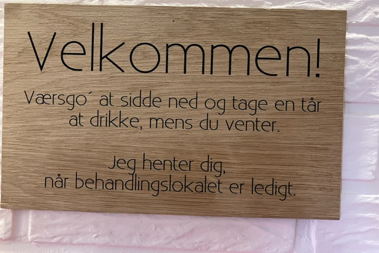 Håndlavet træskilt i klinikken i Løgumkloster: Velkommen! Værsgo at sidde ned og tage en tår at drikke, mens du venter. Jeg henter dig, når behandlingslokalet er ledigt.