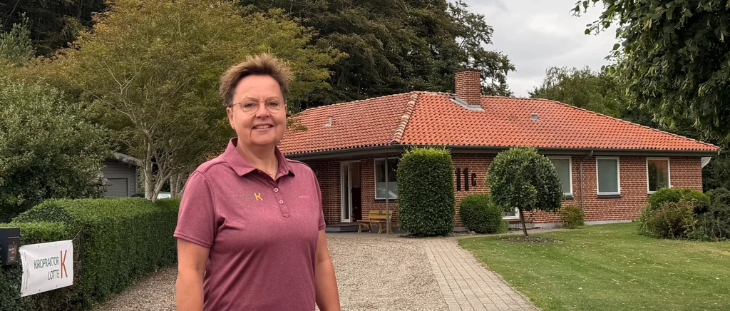 Kiropraktor Lotte Krogh på indkørslen foran klinikken på Sølstedgårdvej 11C i Løgumkloster, med husnummeret 11c på murstensfacaden og klinikkens banner ved hækken