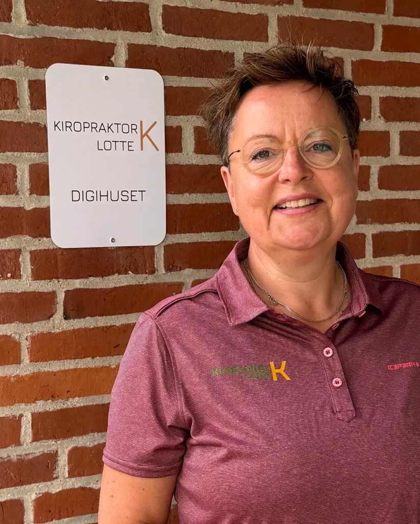 Kiropraktor Lotte Krogh smilende foran klinikskiltet Kiropraktor Lotte K på murstensvæggen ved indgangen til klinikken i Løgumkloster